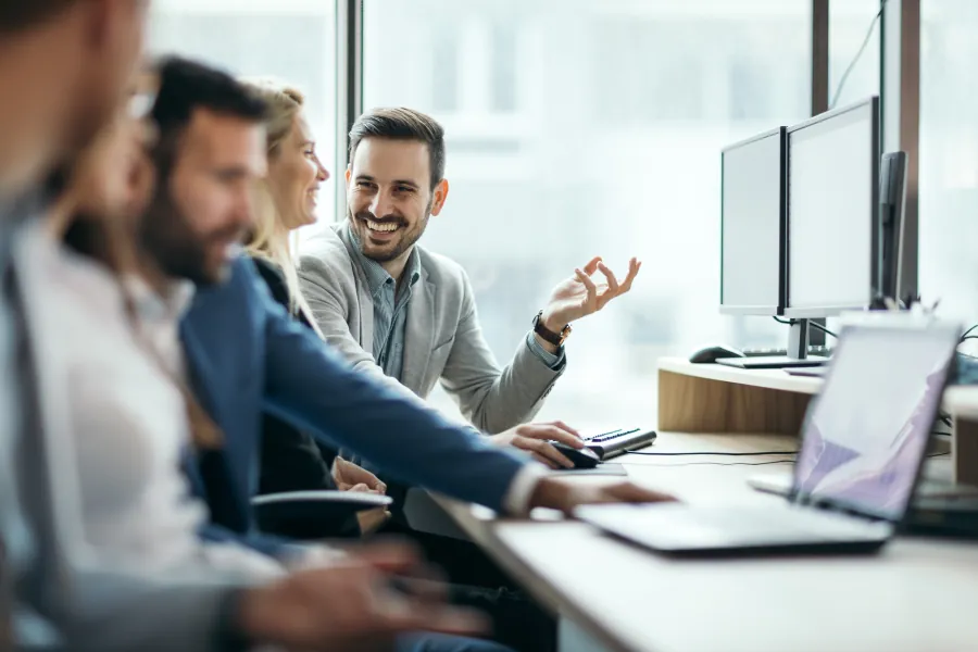 Ein Mann und eine Frau sitzen am Computer und lachen, im Vordergrund sitzen zwei weitere Männer