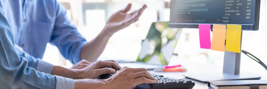 Zwei Männer sitzen am Computer und unterhalten sich und einer von beiden programmiert.