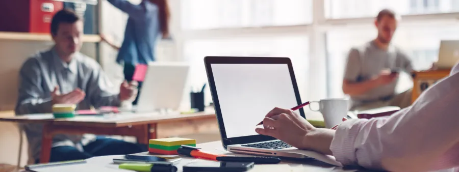 Ein Mann arbeitet an seinem Laptop, im Hintegrund sind weitere Personen und arbeiten 