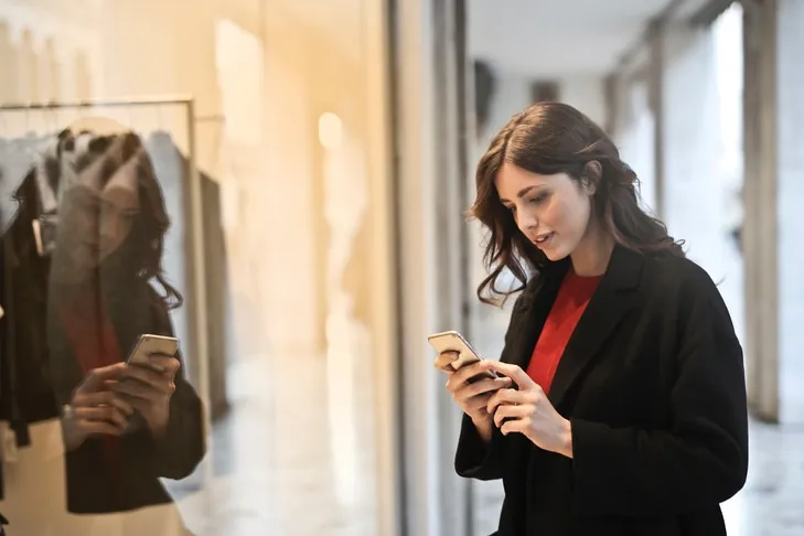 Eine junge Frau schaut auf ihr Smartphone, während sie vor einem Schaufenster steht.